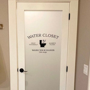 May include: A white bathroom door with a frosted glass panel. The door has a black door handle and a black hook rack with a white towel hanging on it. The glass panel has a black and white sign that reads "WATER CLOSET" with a toilet icon and the text "Seat Yourself 5¢ Always Flush Wash Your Hands Use Soap."