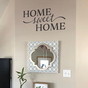 May include: A decorative wall decal with the text "Home Sweet Home" in black lettering. The decal is on a beige wall with a white and blue patterned mirror hanging below it.