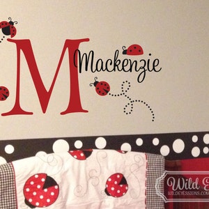 May include: A red letter M with the name Mackenzie written in black cursive script. There are five red ladybugs with black spots and black dotted lines trailing behind them. The background is a light grey wall with a black and white polka dot border.