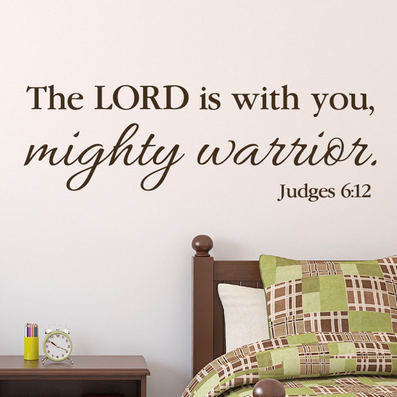 May include: A brown wall decal with the text "The LORD is with you, mighty warrior. Judges 6:12". The decal is placed above a wooden bed frame with a green and brown plaid pillow.