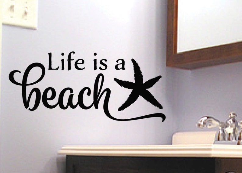 Op de afbeelding: Zwarte en witte wanddecoratie met de tekst "Life is a beach" en een gestileerde zeester.
