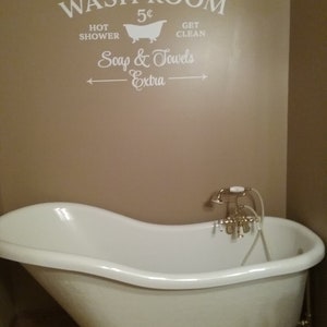 Peut inclure: Une baignoire &agrave; pieds en blanc avec des robinets dor&eacute;s. Le mur derri&egrave;re la baignoire porte une pancarte de style vintage qui indique "Wash Room 5&cent; Hot Shower Get Clean Soap & Towels Extra".