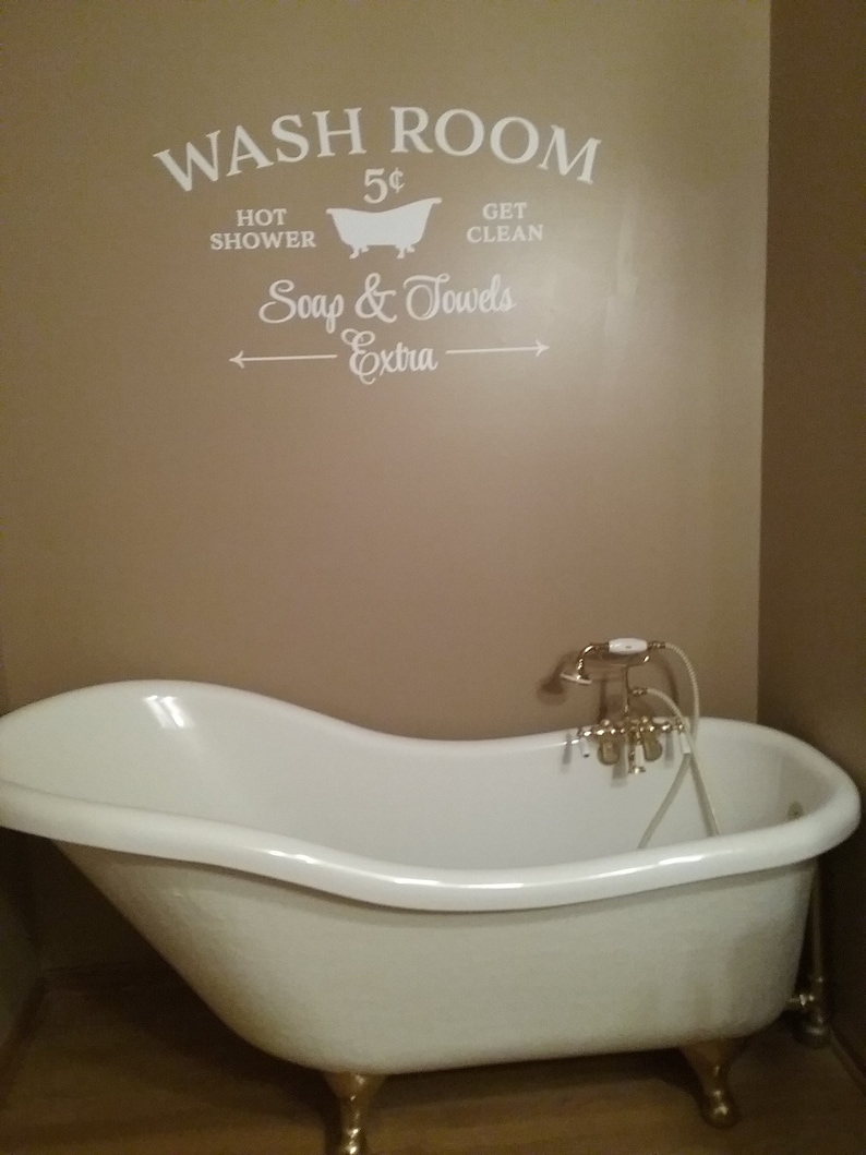 Peut inclure: Une baignoire &agrave; pieds en blanc avec des robinets dor&eacute;s. Le mur derri&egrave;re la baignoire porte une pancarte de style vintage qui indique "Wash Room 5&cent; Hot Shower Get Clean Soap & Towels Extra".