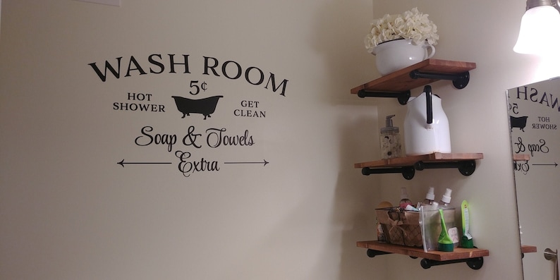 Peut inclure: Une affiche vintage noir et blanc qui dit "WASH ROOM 5&cent; HOT SHOWER GET CLEAN Soap & Towels -Extra-" avec une silhouette de baignoire. L'affiche est sur un mur blanc avec deux &eacute;tag&egrave;res en bois avec des supports en m&eacute;tal noir. Les &eacute;tag&egrave;res sont d&eacute;cor&eacute;es d'un pichet blanc, d'une bouteille en verre, d'un panier et d'un bol en c&eacute;ramique blanc avec des fleurs blanches.