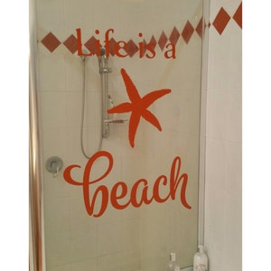 Op de afbeelding: Een douchedeur met een rode vinyl sticker waarop "Life is a beach" staat, met een rode zeester in het midden van de tekst.