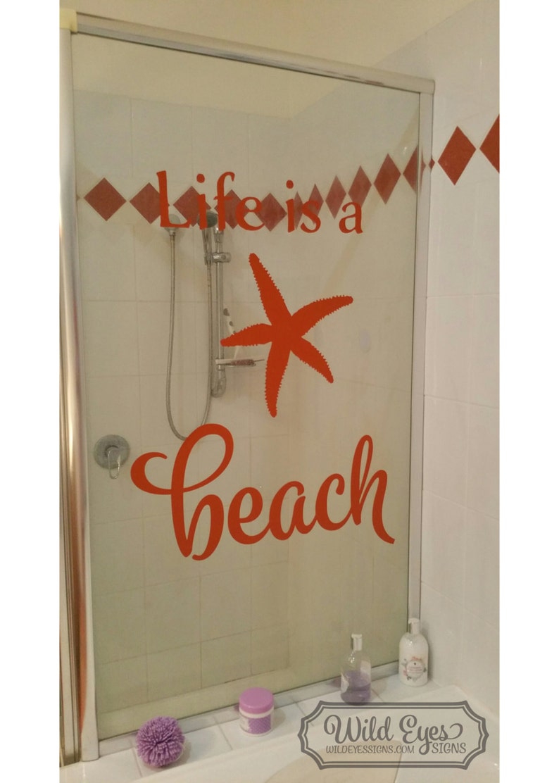 Op de afbeelding: Een douchedeur met een rode vinyl sticker waarop "Life is a beach" staat, met een rode zeester in het midden van de tekst.