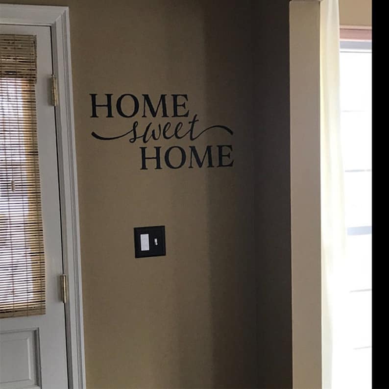 May include: A brown wall with a black vinyl decal that reads "HOME sweet HOME". A white door with a brown bamboo blind is to the left of the decal. A white light switch is to the right of the decal.