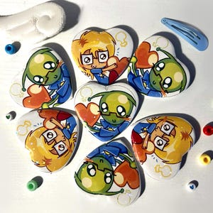 May include: Six heart-shaped badges featuring cartoon illustrations of a green alien and a blonde character with glasses holding orange hearts. The badges are arranged with a white fabric wing, a light blue hair clip, and several small colourful beads.