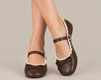 Può includere: Un paio di scarpe basse Mary Jane marrone scuro con punta arrotondata, un cinturino sottile sul collo del piede e un fiocco decorativo sul davanti. Il bordo di ogni scarpa è rifinito con un merletto color crema smerlato e una fibbia.