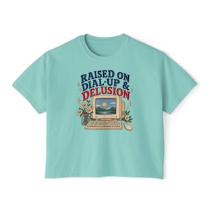 May include: A light teal t-shirt featuring a graphic of a vintage computer with a landscape screen, a keyboard, a mouse, and a vase of daisies. The text reads RAISED ON DIAL-UP & DELUSION in dark blue and red lettering with small yellow star accents.