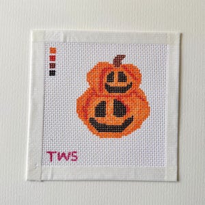 May include: A square needlepoint canvas featuring a cross-stitch design of two stacked orange pumpkins with black facial features. A vertical color key with orange, red, brown, and black squares is in the upper left, and the letters TWS are in pink.