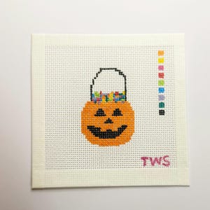 Puede incluir: Un lienzo cuadrado de punto de aguja blanco con un diseño de punto de cruz de un cubo de calabaza naranja lleno de dulces coloridos. A la derecha hay una clave de color vertical con nueve cuadrados pequeños y las letras TWS en rosa.