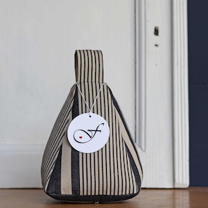 Könnte beinhalten: Ein gestreifter Türstopper in Schwarz und Beige mit Griff. Der Türstopper ist dreieckig und hat ein Etikett mit einem kursiven "f" und einem kleinen roten Herz. Der Türstopper steht auf einer Holzoberfläche.