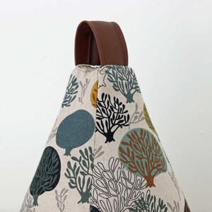 Könnte beinhalten: Ein beiger Türstopper aus Stoff mit einem sich wiederholenden Muster aus stilisierten Bäumen in Grün-, Blau-, Braun- und Schwarztönen. Er hat einen braunen Ledergriff oben. Der Türstopper ist dreieckig geformt.