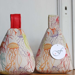 Könnte beinhalten: Zwei dekorative Türstopper mit einem Quallenmuster. Die Türstopper sind kegelförmig und aus Stoff gefertigt. Sie zeigen ein farbenfrohes Quallenmuster in Orange, Lila und Grün auf cremefarbenem Hintergrund. Jeder Stopper hat einen roten Ledergriff.