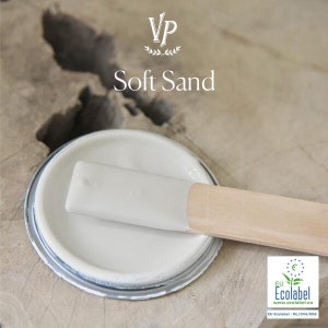 Op de afbeelding: Een gedeeltelijk geopend verfblik met een lichtgrijze verfkleur en een houten roerstaaf. De tekst "Soft Sand" staat boven het blik. Het blik heeft een zilveren rand en staat op een lichtbruin oppervlak.