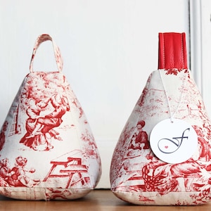 Könnte beinhalten: Zwei kegelförmige Türstopper aus rot-weißem Stoff mit Toile-de-Jouy-Muster. Einer hat einen Stoffgriff, der andere ein rotes Lederoberteil und ein Etikett mit einem stilisierten "f" und einem Herz.