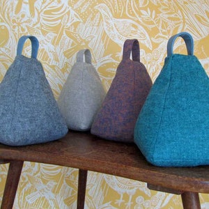 Puede incluir: Cuatro topes de puerta triangulares en tonos gris, morado y turquesa, cada uno con un asa de tela. Est&aacute;n sobre una mesa de madera frente a un papel pintado floral amarillo.