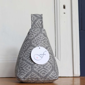 Könnte beinhalten: Ein grauer Türstopper aus Stoff mit geometrischem Muster und Griff. Ein weißes Etikett mit einem stilisierten "f" und einem kleinen rosa Herz hängt am Türstopper. Der Türstopper steht auf einer Holzoberfläche.