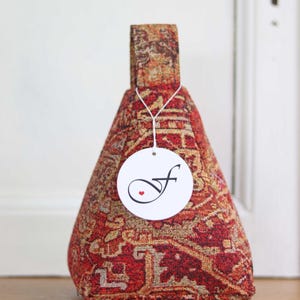 Könnte beinhalten: Dekorativer Türstopper aus rotem und goldenem Stoff mit Muster. Er hat einen Griff und ein Etikett mit einem kursiven "f" und einem kleinen roten Herz. Der Stoff hat ein traditionelles Teppichdesign.