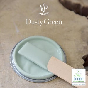 Könnte beinhalten: Eine Dose "Dusty Green" Farbe mit einem Holzrührstab. Die Farbe ist ein blasses Grün, und die Dose hat einen silbernen Rand. Das Bild enthält auch das Ecolabel-Logo.