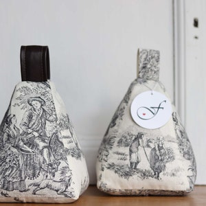 Könnte beinhalten: Zwei dreieckige Türstopper mit Toile-de-Jouy-Muster. Der Stoff zeigt schwarz-weiße Szenen von Menschen und Tieren. Jeder Türstopper hat einen braunen Ledergriff. Sie sind zum Offenhalten von Türen gedacht.