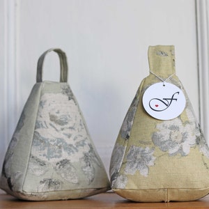 Könnte beinhalten: Zwei dreieckige Türstopper mit Blumenmustern. Einer ist hellgrün mit Griff, der andere gelbgrün mit Etikett. Die Türstopper sind aus Stoff und haben einen abgerundeten Boden.