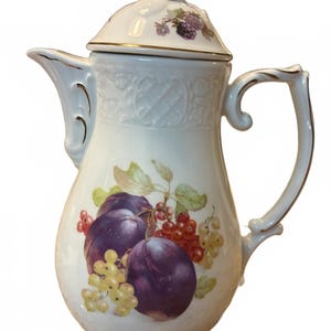 May include: A white porcelain coffee pot featuring a fruit motif with purple plums, red currants, and white berries. The pot has a lid with a small sculpted finial, a curved handle, and a spout, all accented with delicate gold trim along the edges.