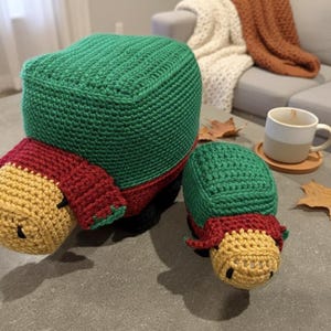 May include: Two handmade crochet turtle toys. The turtles have green shells, red sides, and yellow heads. One turtle is larger than the other. The toys are sitting on a gray surface.