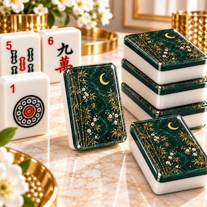 Può includere: Una collezione di tessere del Mahjong composta da tessere bianche con segni numerici e circolari rossi e neri, insieme a tessere verde scuro con disegni dorati di bambù, fiori bianchi e una luna crescente sulla superficie.