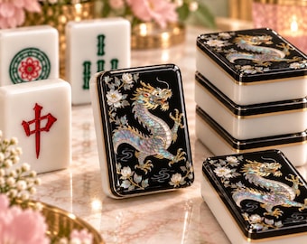 Puede incluir: Un conjunto de fichas de Mahjong con superficies negras, ilustraciones de dragones iridiscentes y detalles florales blancos. Algunas fichas son blancas con símbolos rojos o verdes, mientras que otras están apiladas, mostrando un acabado brillante.