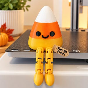 May include: A 3D printed candy corn figurine with a white top, orange middle, and yellow base. It features a black smiling face and articulated yellow legs. One leg holds a small tan tag with the text Fun 3D Prints printed in black.