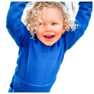 May include: A young child with curly blonde hair wearing a bright blue waffle knit long-sleeve shirt. The child is smiling and looking at the camera.