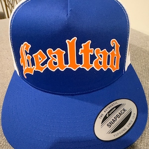May include: A blue and white trucker hat featuring the word Lealtad in orange gothic-style lettering on the front panel. The cap has a flat brim and a circular sticker on the brim that reads The Authentic Yupoong Snapback Since 1974.