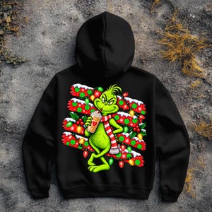 May include: A black hooded sweatshirt featuring a graphic print of a green character wearing a red and white striped scarf, holding a beverage cup with a heart icon, surrounded by red and green Christmas lights and snowy branches.
