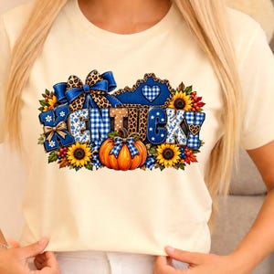 May include: A cream-coloured t-shirt featuring a graphic print of the word KENTUCKY in blue gingham and leopard print letters. The design includes a large orange pumpkin, sunflowers, a blue bow, and a blue state outline with a heart.