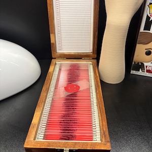 Op de afbeelding: Een houten opbergdoos voor microscoopglaasjes met een scharnierend deksel, voorzien van vijftig genummerde sleuven. Binnenin bevinden zich talloze glazen glaasjes met rood gekleurde preparaten. Eén glaasje met een smiley-icoon.