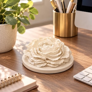 May include: A cream-colored decorative object shaped like a blooming flower with layered, ruffled petals resting on a circular flat base. The sculpture features a textured, matte finish and is positioned as a standalone piece on a flat surface.