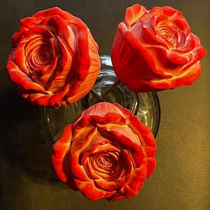 May include: Three red roses with orange and yellow undertones arranged in a clear glass vase. The flowers feature layered, tightly curled petals that create a dense, rounded shape. The stems are visible through the transparent glass container.