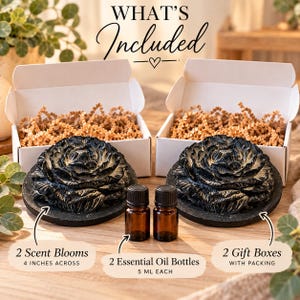 May include: Two black flower-shaped scent blooms measuring 4 inches across, two 5 ml amber glass essential oil bottles, and two white gift boxes filled with brown paper packing material. The text reads WHAT'S Included, 2 Scent Blooms, 2 Essential Oil Bottles, and 2 Gift Boxes WITH PACKING.