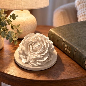 May include: A white, sculpted, three-dimensional flower ornament resting on a circular white base. The flower features intricate, layered petals with a matte finish. Beside the ornament lies a dark green, leather-bound Holy Bible book.