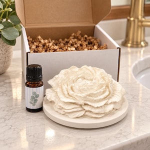 May include: A white ceramic flower diffuser on a circular base, accompanied by a small amber glass bottle with a black cap and a label featuring a blue striped pattern and a green leaf illustration, all presented in an open white cardboard box.
