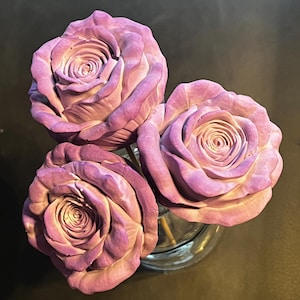 May include: Three sculpted rose flowers with a matte finish in shades of light purple and lavender. Each rose features layered, textured petals arranged in a circular pattern, attached to thin stems held within a clear glass container.
