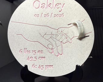 Può includere: Una targa circolare color bianco sporco maculato con un'incisione che raffigura una mano adulta che tiene la mano di un neonato. Il testo riporta Oakley, 02 / 26 / 2026, 6 lbs 15 oz, 49,5 cm e 6:45 pm. È attaccato un piccolo braccialetto identificativo ospedaliero bianco.