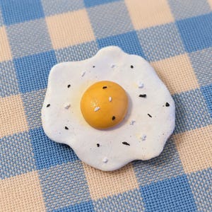 May include: A sculpted fried egg pin featuring a smooth, rounded yellow yolk centered on an irregular, matte white egg white. The surface is speckled with small, scattered black and white flecks, mimicking the appearance of pepper and salt.