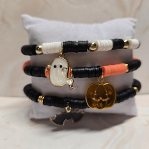 May include: A set of three stretch bracelets featuring black, white, and orange clay disc beads with gold-tone spacers. The bracelets include dangling charms shaped like a white ghost, an orange pumpkin, and a black bat with gold-tone accents.