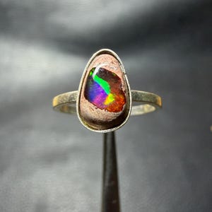 Puede incluir: Un anillo de plata con un ópalo boulder en forma de lágrima engastado en un bisel de metal liso. El ópalo muestra un vibrante juego de colores con destellos verde neón, azul eléctrico y morado intenso sobre una matriz marrón texturizada.