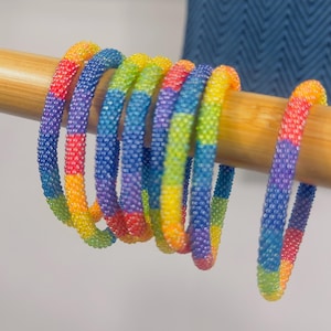Puede incluir: Una colección de pulseras circulares hechas de pequeñas cuentas de vidrio multicolores dispuestas en un patrón de arcoíris repetitivo de rojo, naranja, amarillo, verde, azul y morado. Las pulseras se muestran colgadas de una varilla de madera clara.