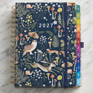 May include: A 2027 spiral-bound planner with a dark blue cover featuring a pattern of mushrooms, ferns, flowers, a moth, an owl, a squirrel, a hare, and snails. The planner includes a vertical elastic closure and colorful monthly index tabs.