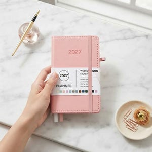 May include: A light pink 2027 weekly and monthly planner with a matching elastic band closure. The cover features the year 2027 embossed in the center. A white paper band wraps around the middle, displaying the text 2027 PLANNER.